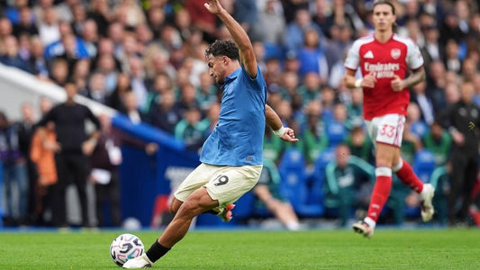Kostoulas lövés közben a Brighton Arsenal elleni 3–0-s győzelmén, piros-fehér Arsenal-játékossal a háttérben